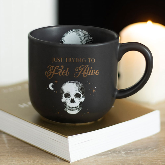 A black mug with a white skull pattern inside and the words Just Trying To Feel Alive in gold written on the outside with white skull, moon and stars showing everyone how your day is going while you enjoy a nice warm drink.