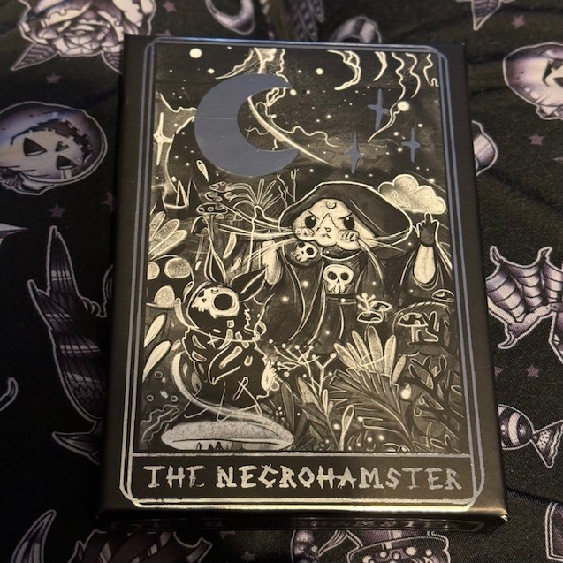 The Necrohamster card game front of the box. The box is black with silver writing and imagery representing a necromancer hamster, undead bunny and crescent moon