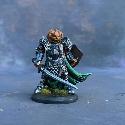 A Reaper Miniature Halloween Knight hand painted by Mrs MLG, a fierce pumpkin headed warrior wearing armour, holding a sword in one hand and a coffin lid shield in the other with a green cape flapping behind. 