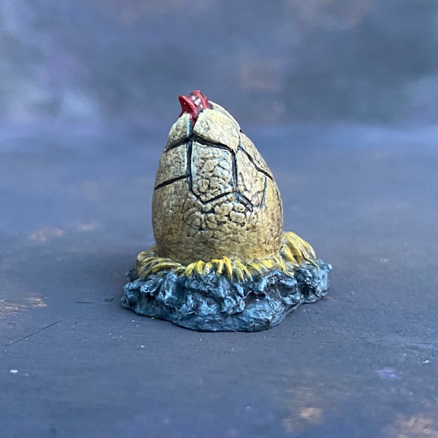 A Mantic Games objective marker in the style of a baby red dragon hatching from its egg. This cute little miniature has a red dragon snout, cracked egg on a bed of hay and stones.