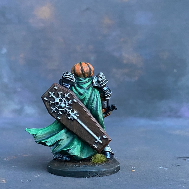 A Reaper Miniature Halloween Knight hand painted by Mrs MLG, a fierce pumpkin headed warrior wearing armour, holding a sword in one hand and a coffin lid shield in the other with a green cape flapping behind. 