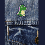 An extremely cute little enamel pin badge of a green dinosaur with pin spikes down its back and a yellow tummy. The badge on some jeans